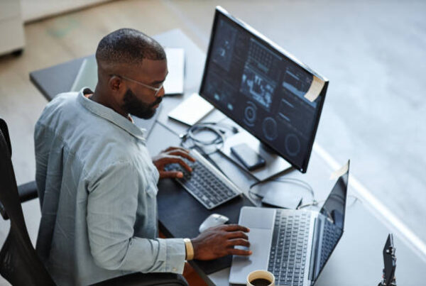Black male IT professional analyzing data on computer and laptop in a modern workspace